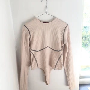 Ribbad Långärmad Bodysuit - En beige ribbad långärmad bodysuit från Boohoo med sömmar i kontrast. Endast använd en gång. Köpt för 252 kr.