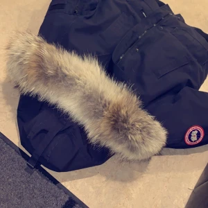 Chilliwack bomberjacka Navy - Säljer en varm dunjacka i färgen navy i bombermodell med pälsprydd luva från Canada Goose i ett slitstarkt material. Denna bomberjacka stängs med både dold dragkedja och knappar och den formbara luvan går att reglera. Framtill finns flera fickor med skön insida i fleece. Rejäla ribbstickade muddar i ärmslut och nederkant utan skav. Flera bilder kan skickas och självkart kan äktheten styrkas. Mvh Lillia&Kayn