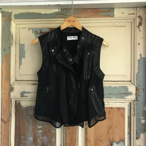  - Fin väst i svart konstskinn i romantisk stil med det skira svarta tyget på utsidan. Lika snygg till en färgstark klänning som till ett par svarta byxor! 