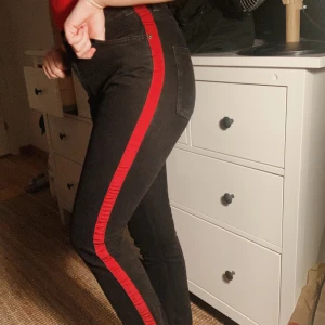 SVARTA JEANS, MONKI, STRL 27 - Svarta jeans med röd rand på båda yttersidorna av benen. Storlek 27,  jag är 170cm och väger 64kg. Går lite längre än ankellängd på mig när jag står upp!