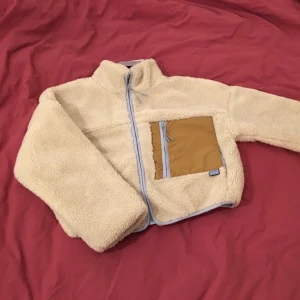 Mysig teddyjacka från Pull & Bear - En vit/beige jacka från Pull & Bear med ljusblå detaljer och en stor fick fram. Oanvänd med prislapp kvar. Storlek L.
