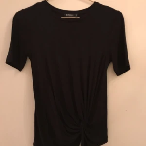  - Svart topp från Tiger AW17 i storlek XS med drapering, kort ärm och snyggt fall. Rund hals med knut framtill. 