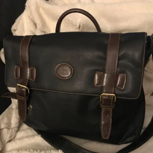  - Svart väska med bruna detaljer. Längd 36 cm, höjd 30 cm och bredd 10 cm. Dragkedja. Justerbar axelrem. Svart foder. En blixtlås försedd innerfack. Använd några fåtal gånger. Ser helt ny ut. Köparen betalar frakt (ca 60 kr)