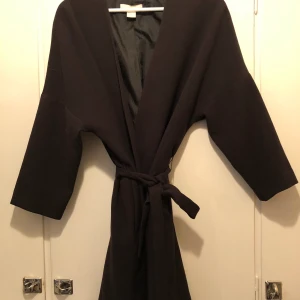 Svart kappa med skärp i midjan  - Svart kappa i mjukt material från HM. Skärp/band i midjan. Storlek S. Använd ett fåtal gånger. Nypris 499:-.