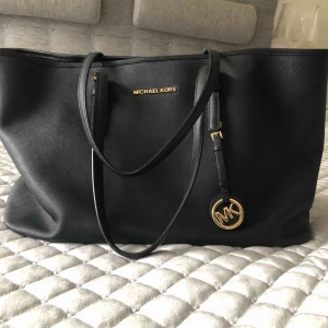 - Användbar Jet set travel väska från Michael Kors. Såklart äkta! Svart läder med gullddetaljer. 40 cm lång, 15 cm bred och 26 cm hög/djup. Lite över 2 år gammal men sparsamt använd. Inte alls använd på över 1 år. Förvaringspåse finns och medföljer. 