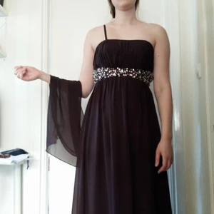  - Balklänning/Aftonklänning i strl M enligt lappen som fortfarande sitter kvar.  Varmbrun, med avtagbara axelband och sjal i samma tyg som klänningen. Extra fint ryggparti. Två lager i kjolen som är lång nog att vara balklänning för en 180 lång utan klackskor.  Säljer på grund av för stor. 