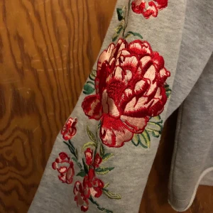 Hoodie med broderi - Grå hoodie med broderade blommor på ärmarna och på luvan! Supersöt. Lite cropped där fram. 