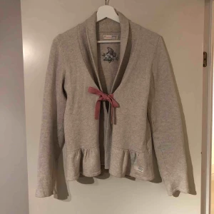  - Stickat kofta från Odd Molly i en fin beige färg med knytning fram i rosa. Fina volang detaljer fram och ganska djup urringning. Fin feminin passform och i fint skick, passformen är ganska tight. Strl 3 = M/L 