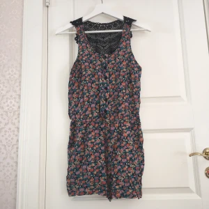  - Blommig (lila, grön & orange) shortsdress, svart virkad bak, stängs med knappar och fickor på sidorna. 