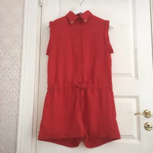  - Orange/röd shortsdress med band, stängs med knappar, kragen har gulddetaljer och låtsas fickor på baksidan. 