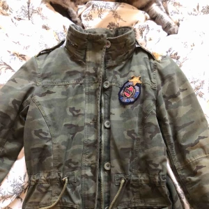  - Jättefin höst/vår camo-jacka med guldpaljetter på axlarna. Inköpt ca 6 år sedan för runt 4000 och aldrig använd. Det finns en luva man kan vecka ut från halsen Och man kan även dra åt så den blir tightare i midjan. I perfekt skick då den är oanvänd