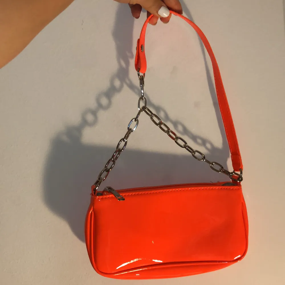 Liten neon-orange färgad baguette stil handväska. Lackat material med silver detaljer. Handtag avtagbar på ena sidan. Helt oanvänd. Köparen står för frakt ifall man vill ha det fraktat. . Väskor.