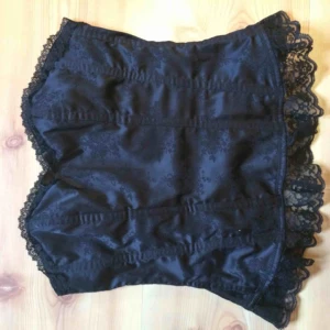 Beauty - Korsett i fint skick! Svart siden med blomstermönster. Spets kring kanter och en liten rosett vid bysten. Saknar storlekslapp men uppskattar den till XS-S, kupan inte större än B. 