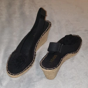 Svarta klackskor med flätad kilklack och öppen tå - Aldrig använda. Blom-liknande dekoration vid tån. Använder strumpor för hygienskäl. Storlek 40 men passar 39 bättre