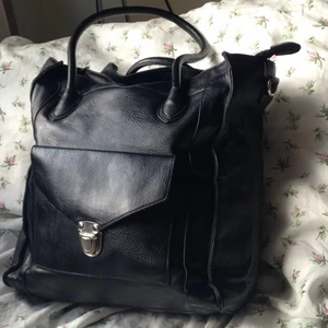  - Svart handväska i faux läder.  Snygga detaljer. Silverfärgat spänne på facket på framsidan. Mycket rymlig. Innerfack med dragkedja. Tillhörande axelrem som går att haka fast (se bild 2). Dragkedja till stora facket (se bild 2).  Höjd: 33 cm Bredd: 29 cm Djup: 15,5 cm