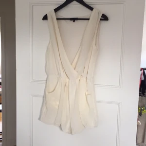  - Byxdress i en fin cremevit färg. Omlott framtill och medföljer ett band att knyta vid höften. Går även att använda bak-och-fram så ryggen blir djup istället. Fickor på sidorna. Aldrig använd. 