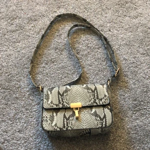 Väska från glitter - Ormmönstrad väska från glitter. Den är i bra skick och har ett axelband som man både kan ha kort och långt. Det finns en liten ficka inuti väskan med dragkedja. Väskans knäppe är i guld