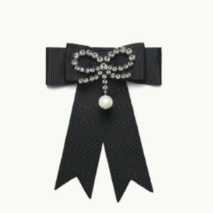  - ERDEM x H&M. En brosch i metall med ripsrosett. Rosetten är dekorerad med en mindre rosett i strass samt en pärlemorskimrande pärla i plast. Bredd 10,5 cm, längd 11,5 cm. Broschen levereras i en blommönstrad pappask med lock. Locket hålls samman med ripsband.