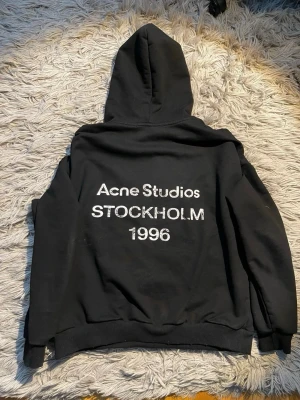 Acne Studios hoodie - Svart hoodie från Acne Studios med tryck på ryggen där det står 'Acne Studios STOCKHOLM 1996' i vitt. Klassisk passform med huva och ribbade muddar vid ärmslut och nederkant. Tillverkad i mjukt bomullsmaterial för en bekväm känsla. Perfekt hoodie nu till våren! Bara att höra av sig vid minsta funderingar!