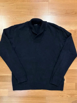 Svart stickad tröja med krage - Mörkblå stickad tröja från R. Lauren med bred ribbad krage och muddar vid ärmslut och nederkant. Tröjan har en klassisk, avslappnad passform och är perfekt för kyliga dagar. Materialet känns mjukt och bekvämt mot huden.