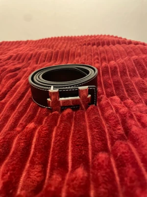 Svart skärp med silverspänne - Stilrent svart bälte med ett stort, blankt silverspänne i form av ett H. Skärpet har vita kontrastsömmar längs kanterna och är gjort i slätt material som ger en klassisk look. Perfekt accessoar för att lyfta din outfit.