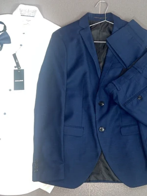 Marinblå kostym från Jack & Jones - Snygg marinblå kostym från Jack & Jones med klassisk kavaj och matchande byxor. Kavajen har två knappar, smala slag och fickor framtill. Kostymen är tillverkad i ett slätt material och har en stilren look som passar perfekt till skjorta och slips.