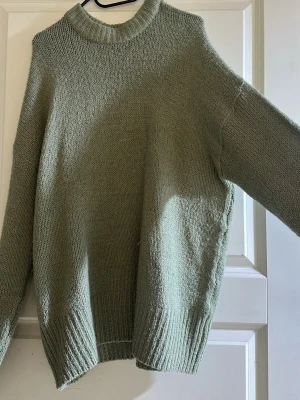 Olivgrön stickad oversized tröja - Mysig stickad tröja i olivgrönt med avslappnad, oversized passform. Tröjan har rund halsringning, långa ärmar och breda ribbade muddar vid ärmslut och nederkant. Perfekt för lager-på-lager och chill dagar.