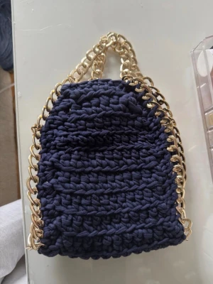 Mörkblå virkad handväska med guldkedja - Unik handväska i mörkblått, grovt virkat tyg med detaljer i form av en tjock guldfärgad kedja som handtag och kant. Väskan har en mjuk, flexibel form och öppet huvudfack. Perfekt för dig som gillar trendiga accessoarer.