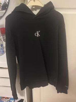 Svart hoodie från Calvin Klein Jeans - Svart hoodie från Calvin Klein Jeans med klassisk huva och vit cK-logga på bröstet. Mjuk bomullskänsla och ribbade muddar vid ärmslut och nederkant. Enkel och stilren design som passar perfekt till jeans eller joggers.