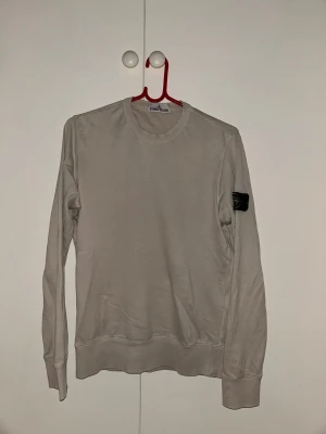 Beige sweatshirt från Stone Island - Säljer en stilren beige sweatshirt från Stone Island med klassisk rund hals och ribbade muddar. Tröjan har den ikoniska svarta loggan på vänster ärm. Perfekt för en avslappnad och trendig look.