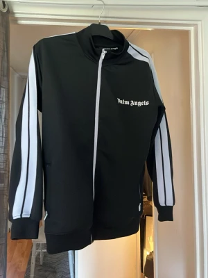 Svart track jacket från Palm Angels - Säljer en svart track jacket från Palm Angels med vita ränder längs ärmarna och vit dragkedja. Jackan har hög krage, två fickor med dragkedja och logga på bröstet. Perfekt för en sportig och trendig look.
