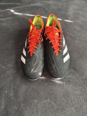 Adidas fotbollsskor svarta med röda detaljer - Säljer ett par snygga fotbollsskor från Adidas i svart med röda snören och gula insida. Skorna har klassiska vita ränder på sidorna och är tillverkade i syntetmaterial som ger bra grepp och komfort på planen. Elite modell