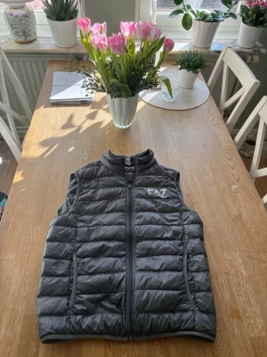 Emporio Armani väst - Snygg svart dunväst från EA7 Emporio Armani med quiltad design och dragkedja framtill. Västen har hög krage och logotyp på bröstet. Perfekt för lager-på-lager och ger en sportig look. Materialet är lätt och isolerande.