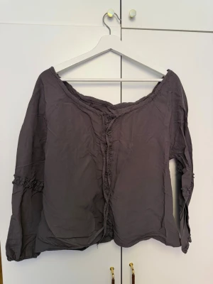 Mörkgrå offshoulder blus - Snygg mörkgrå offshoulder blus med långa ärmar och volangdetaljer längs ärmslut och framkant. Blusen har en avslappnad passform och är tillverkad i ett lätt, skrynkligt tyg som ger en bohemisk känsla.