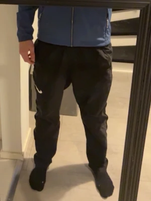 Svarta Nike mjukisbyxor - Säljer ett par svarta mjukisbyxor från Nike med normal passform och klassisk vit logga på benet. Perfekta för chill eller träning, med elastisk midja och bekväm känsla. Passar dig som vill ha en enkel och stilren look. Lite slitna