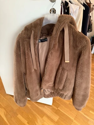 Brun teddyjacka med huva från Zara - Mysig brun teddyjacka från Zara med dragkedja, huva och breda snören. Jackan har två fickor framtill och är fodrad på insidan av luvan. Perfekt för kalla dagar när du vill vara både varm och snygg. Materialet är fluffigt och ger en trendig look. Bra skick! 