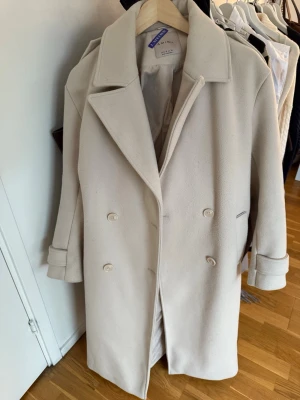 Beige kappa  - Stilren beige kappa med dubbelknäppning, bred krage och bälte i midjan. Kappan har en rak passform och släta fickor på sidorna. Perfekt för dig som gillar minimalistisk och klassisk stil. Kappan har två hål inifrån men går att sy till utan problem. Annars är den i hyfsat skick 