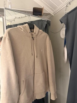 Beige hoodie från Ralph Lauren - Snygg beige hoodie från Ralph Lauren med dragkedja framtill och klassisk huva med dragsko. Har fickor på sidorna och den ikoniska vita loggan broderad på bröstet. Tillverkad i mjukt bomullsmaterial som känns riktigt skönt mot huden.