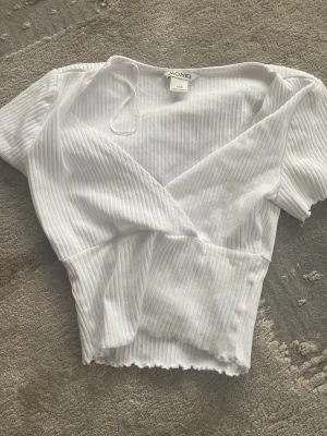 Vit omlottopp från Monki - Säljer en vit omlottopp från Monki i ribbad bomull. Toppen har korta ärmar, v-ringning och croppad passform med vågig kant nertill. Perfekt för dig som gillar en enkel men trendig stil. Använd typ 1 -2 gånger🫰🏼🫰🏼💕💕