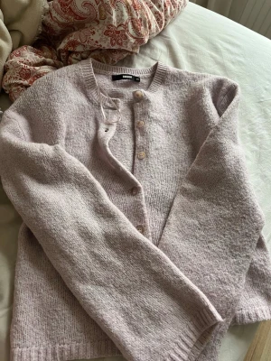 Ljusrosa kofta från BikBok - Mysig ljuslila kofta från BikBok med rund halsringning och knäppning framtill. Koftan är stickad i ett mjukt material och har långa ärmar samt ribbade muddar vid ärmslut och nederkant. Perfekt för lager-på-lager-stil.