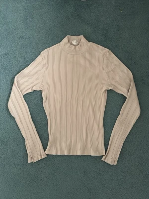 Beige ribbad polotröja - Säljer en beige ribbad polotröja med lång ärm och smal passform. Tröjan har en halv polo-krage och är tillverkad i ett stretchigt material som sitter snyggt på kroppen. Perfekt att styla med jeans eller kjol. Storlek L men passar lika bra på folk med storlek M!