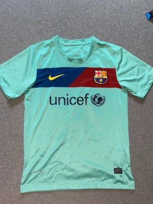 Turkos FC Barcelona matchtröja Nike - Snygg turkos FC Barcelona matchtröja från Nike med klubbmärke och Unicef-logga på bröstet. Tröjan har en diagonal blå och vinröd rand över bröstet samt gul Nike-swoosh. Tillverkad i lätt och ventilerande material, perfekt för fotbollsträning eller match. Passar S-M