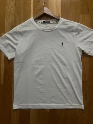 Vit t-shirt från Polo Ralph Lauren - Klassisk vit t-shirt från Polo Ralph Lauren med rund halsringning och den ikoniska lilla marinblå logon broderad på bröstet. Tillverkad i mjuk bomull och har en normal passform med korta ärmar. Perfekt basplagg till garderoben.