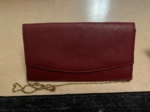 Vinröd kuvertväska med guldkedja - Stilren vinröd kuvertväska med elegant guldfärgad kedja. Väskan har magnetlås och insidan är fodrad med ett brunt tyg med blommönster. Perfekt för dig som vill ha en enkel men snygg accessoar till din outfit.