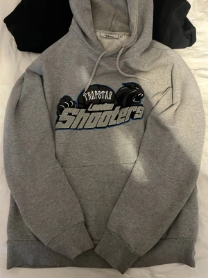 Grå Trapstar London Shooters hoodie - Säljer en grå hoodie från Trapstar med fet 'London Shooters'-logga och svart pantermotiv på bröstet. Hoodien har huva och stor magficka. Tillverkad i mjukt sweatshirtmaterial, perfekt för chill eller streetwear. Stor i storleken 