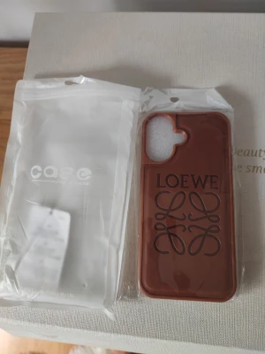 Brunt Loewe mobilskal i läderlook - Snyggt brunt mobilskal från Loewe med elegant logga och mönster på baksidan. Skalet har en stilren design och är gjort i material som liknar läder. Passar perfekt för dig som vill ha ett exklusivt och trendigt skydd till din mobil.
