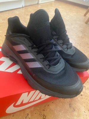 Svarta Adidas sneakers med lila ränder - Snygga svarta sneakers från Adidas med klassiska tre ränder i lila på sidorna. Ovandelen är i mesh och syntetmaterial för bra andningsförmåga och komfort. Skorna har svarta snören och en sportig, låg profil som passar till många olika stilar.