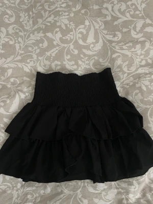 Svart volangkjol från Neo Noir - Supersnygg svart kjol från Neo Noir med bred smock i midjan och dubbla volanger. Kjolen har en kort, flowy passform och är gjord i ett lätt tyg som ger en luftig känsla. 