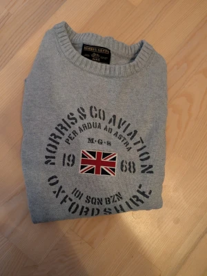Grå tröja från Morris med tryck - Grå långärmad tröja från Morris med rund halsringning och tryck framtill med brittisk flagga och texten 'Morris & Co Aviation Oxfordshire'. Storlek L, fint skick.