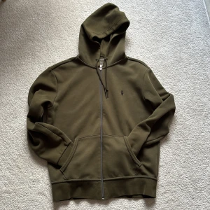 Olivgrön hoodie från Polo Ralph Lauren - Snygg olivgrön hoodie från Polo Ralph Lauren med dragkedja och huva. Tröjan har snörning vid huvan, två fickor fram och den klassiska Polo-loggan broderad på bröstet. Perfekt för chill dagar och enkel att matcha med jeans.
